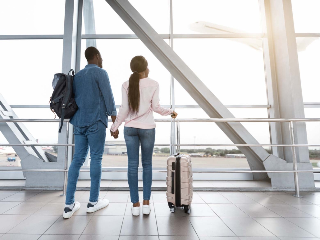 Couple watching planes at airport window with luggage – visitors insurance Canada